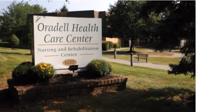Outdoor view of a sign for Oradell Health Care Center, a nursing and rehabilitation center, surrounded by grass, bushes, and trees with a building and benches visible in the background.
