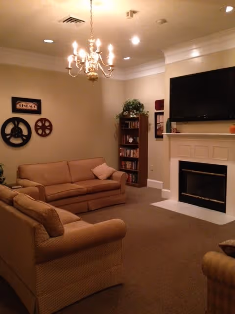 A cozy living room with beige sofas arranged around a fireplace with a mounted flat-screen TV above it. There is a chandelier hanging from the ceiling, a bookshelf filled with books and a plant on top, and decorative wall art including film reels and a framed sign.
