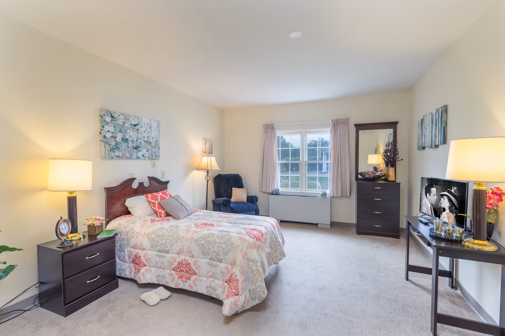 A bright and cozy bedroom in an assisted living facility featuring a bed with patterned bedding, a nightstand with a lamp and clock, a comfortable armchair, a dresser with a mirror, a window with curtains, and a TV on a stand playing a black and white movie.
