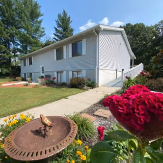 Exterior view of a two-story residential building with light gray siding, surrounded by a well-maintained lawn and garden with colorful flowers. A concrete walkway leads to the entrance, and there are tall trees in the background under a clear blue sky.