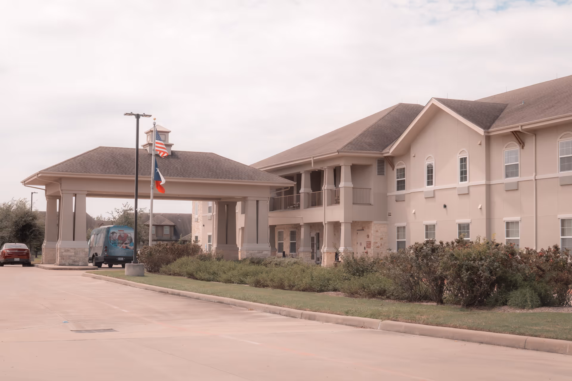 Exterior view of The Legacy at Falcon Point senior living facility showing a covered entrance with two flagpoles flying the American and Texas flags, a driveway, and a beige two-story building with multiple windows and a balcony.