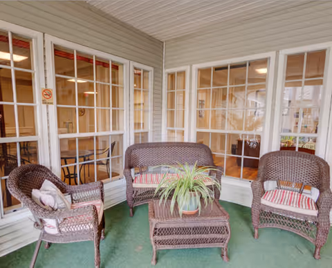 A cozy indoor patio area with three brown wicker chairs and a matching wicker coffee table with a potted plant on it. The chairs have red and white striped cushions. The patio is enclosed with large windows looking into other interior rooms of the facility.