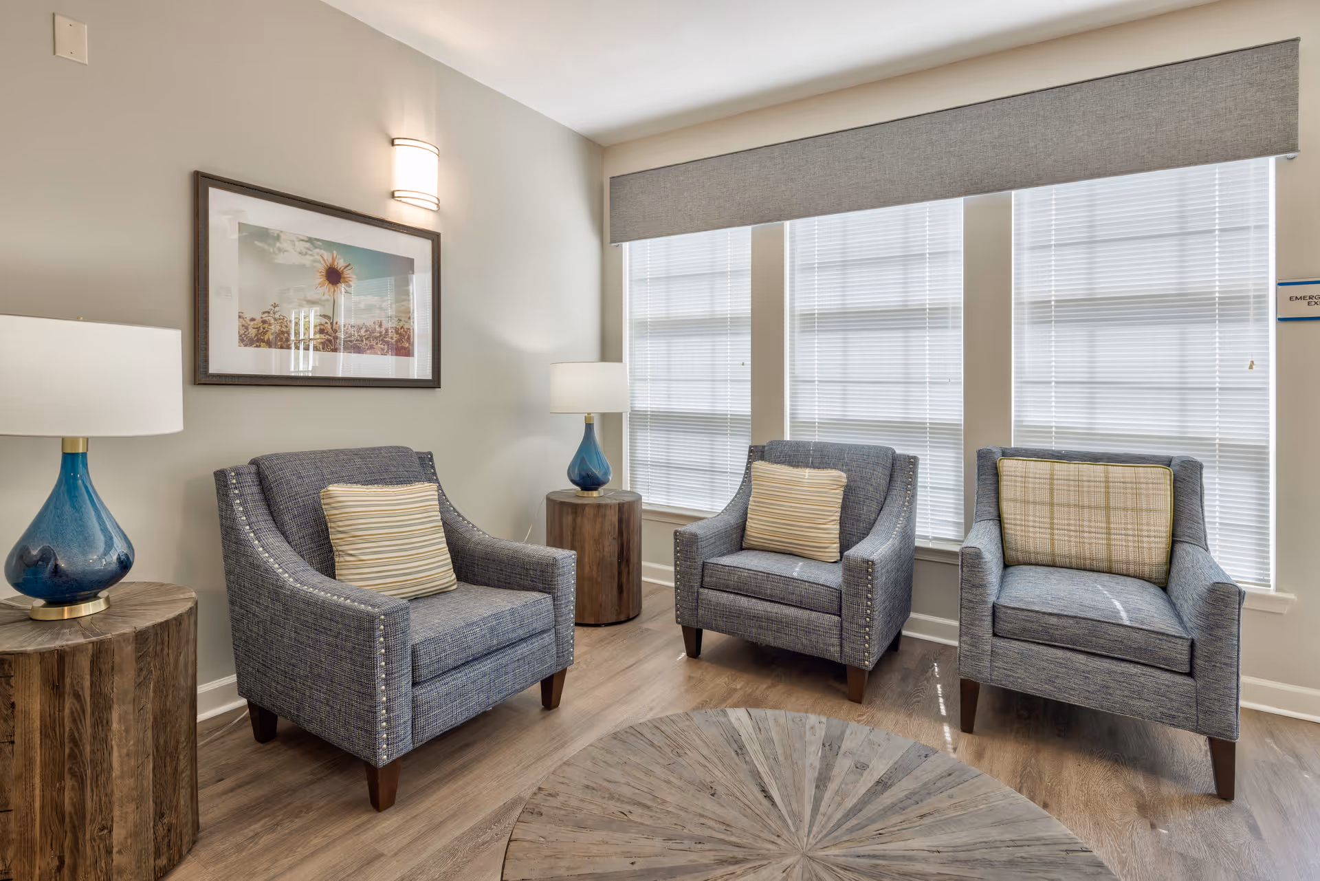 A cozy sitting area with three gray upholstered armchairs, each with a decorative pillow, arranged around a round wooden coffee table. Two wooden side tables with blue glass lamps flank the chairs. The room has light-colored walls, large windows with blinds, and a framed picture of a sunflower on the wall.