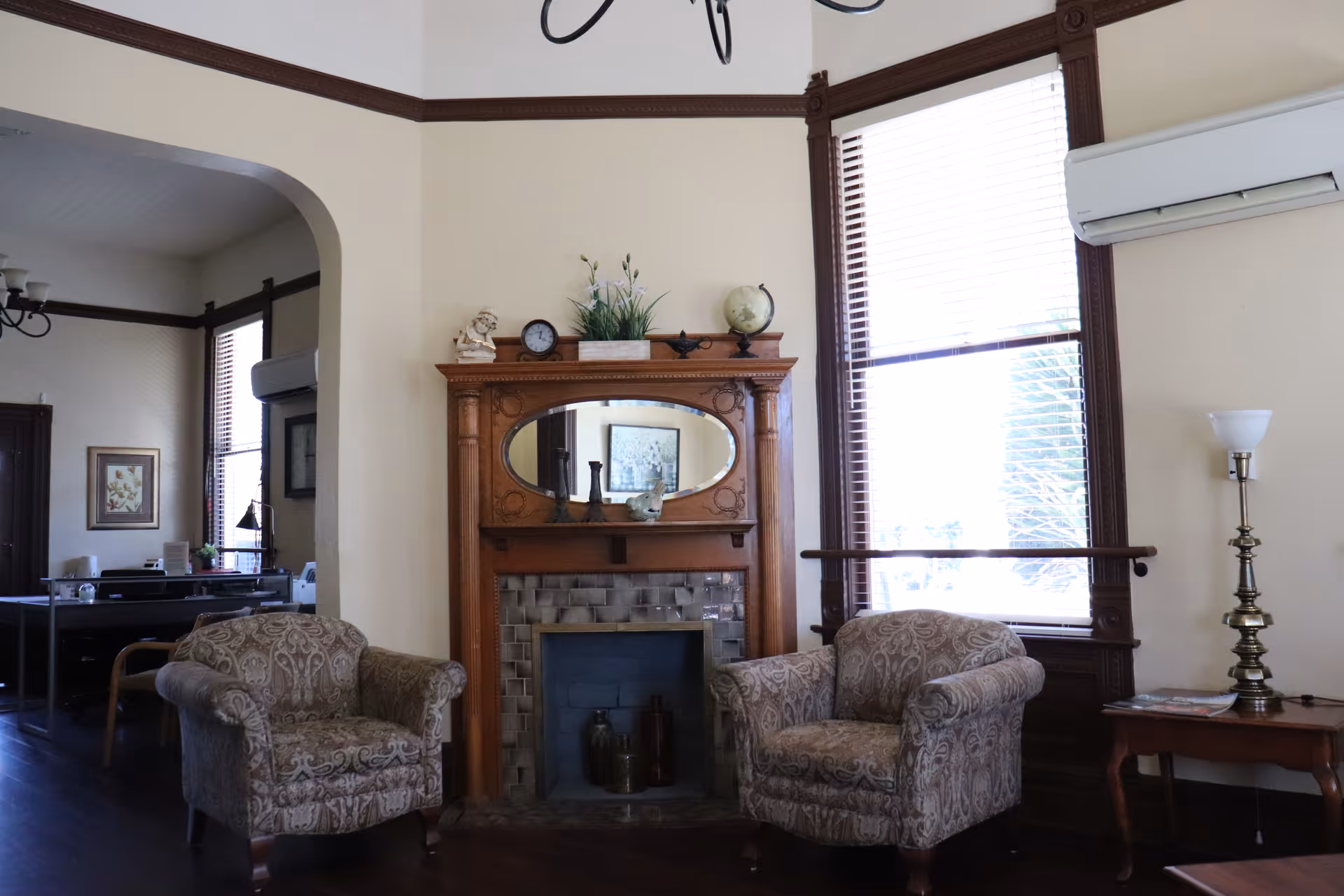 A cozy living room area with two patterned armchairs facing a decorative wooden fireplace with an oval mirror above it. The fireplace mantel is adorned with small decorative items including a clock, a globe, and a small statue. To the right, there is a window with wooden blinds and a tall floor lamp next to a small wooden side table. The room has cream-colored walls with dark wood trim and dark hardwood flooring. In the background, an adjacent room with a desk and framed artwork is visible through an arched doorway.