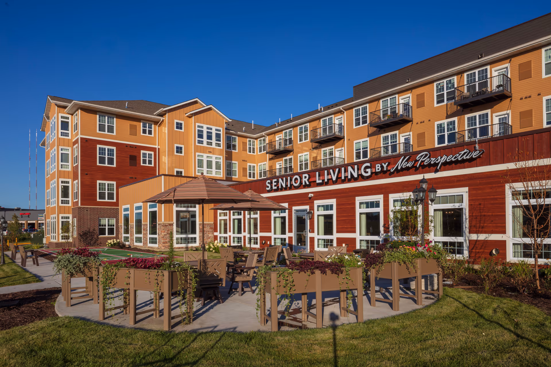 Front exterior of New Perspective Senior Living building featuring an outdoor patio with tables, umbrellas, planters, and a large 'Senior Living' sign.