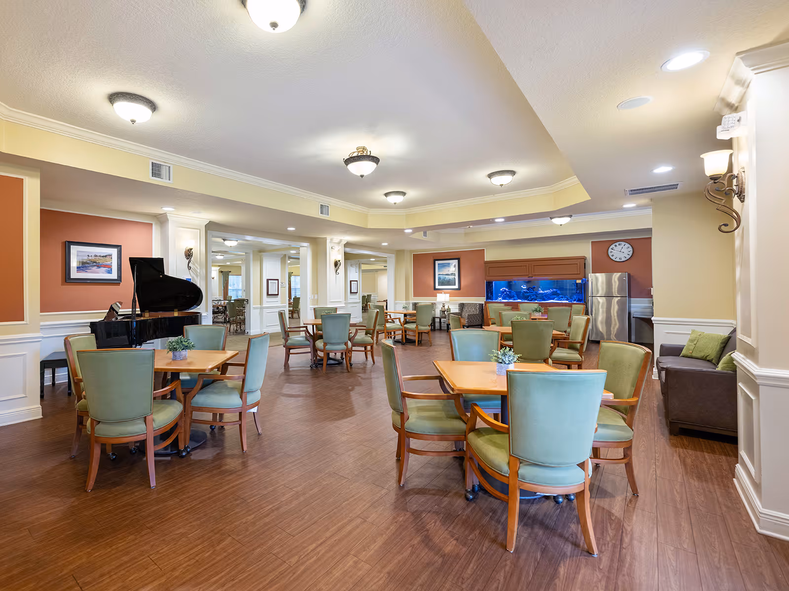 A spacious common area with multiple wooden tables and green cushioned chairs arranged throughout the room. There is a grand piano on the left side, a large aquarium with blue lighting on the back wall, a stainless steel refrigerator, and a clock above it. The room has warm lighting with ceiling fixtures and wall sconces, wood flooring, and light-colored walls with white wainscoting.