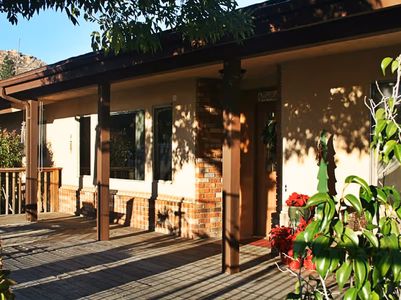 Exterior view of a single-story building with a covered wooden porch supported by brown pillars. The building has beige walls with brick accents around the windows and door. There are plants and flowers near the entrance, and shadows of tree branches are cast on the walls and porch floor.