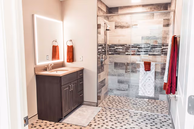 A modern bathroom featuring a dark wood vanity with a sink and illuminated mirror above it. The shower area has glass sliding doors, tiled walls with a decorative horizontal strip, and a pebble-patterned floor. Red and white towels hang on towel rings and hooks on the walls.