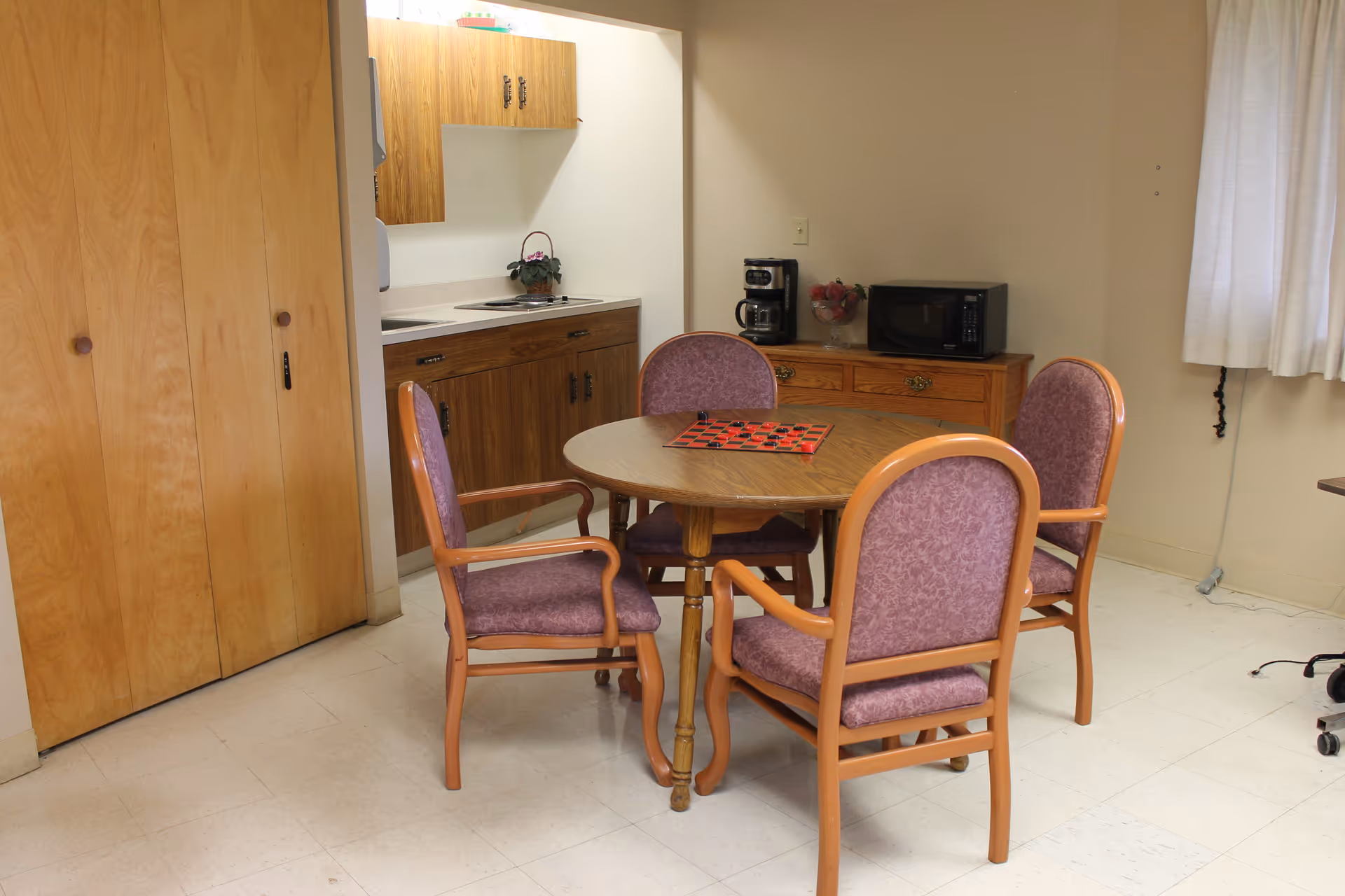 A small dining area with a round wooden table surrounded by four purple upholstered chairs with wooden frames. On the table is a checkers game board. In the background, there is a kitchenette with wooden cabinets, a coffee maker, a microwave, and a small plant. The room has light-colored walls and a window with white curtains.