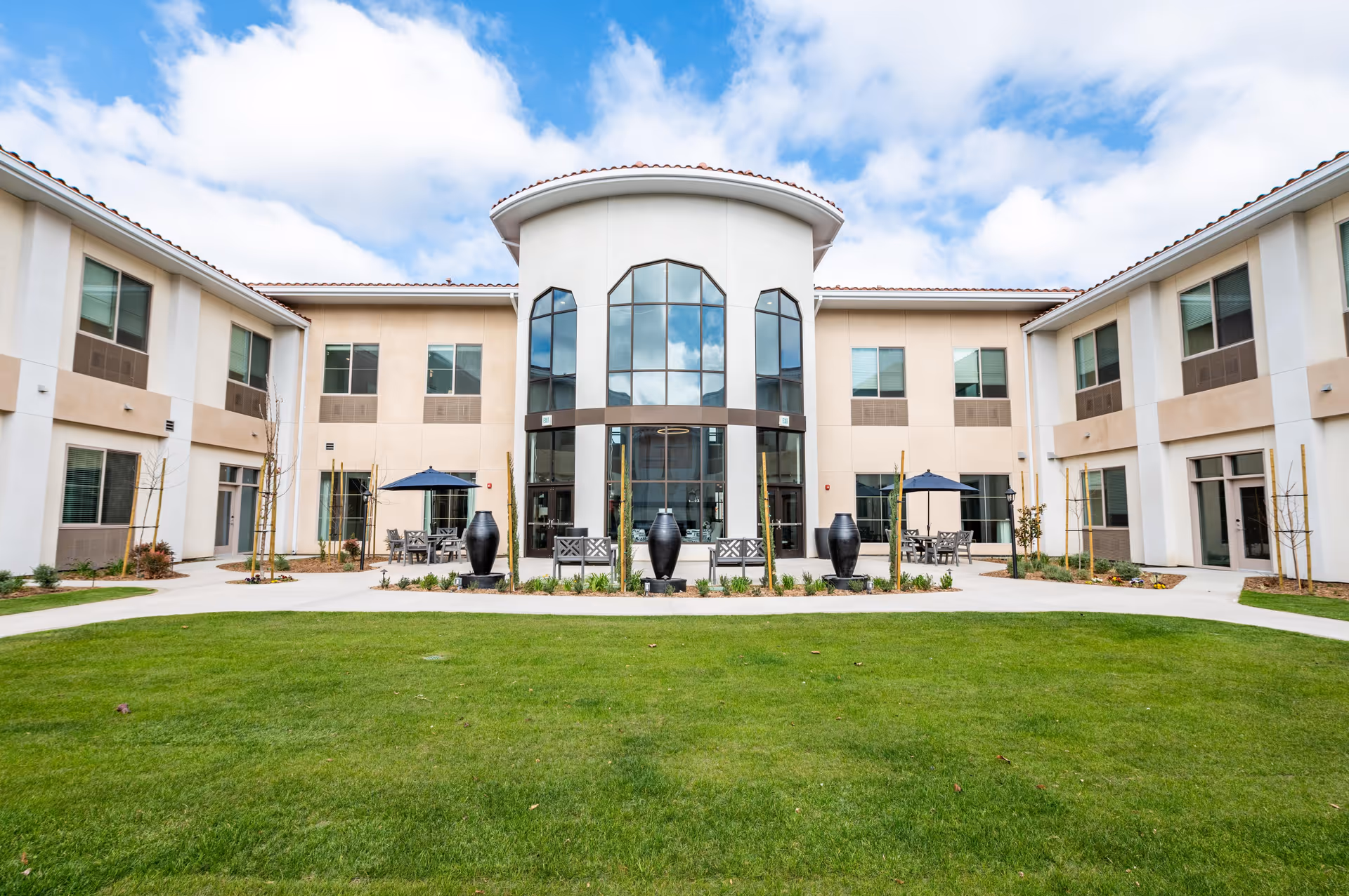 Exterior view of a two-story assisted living facility with a central rounded section featuring large windows. The building surrounds a green lawn with paved walkways, outdoor seating areas with tables and umbrellas, and decorative black vases. The sky is partly cloudy.