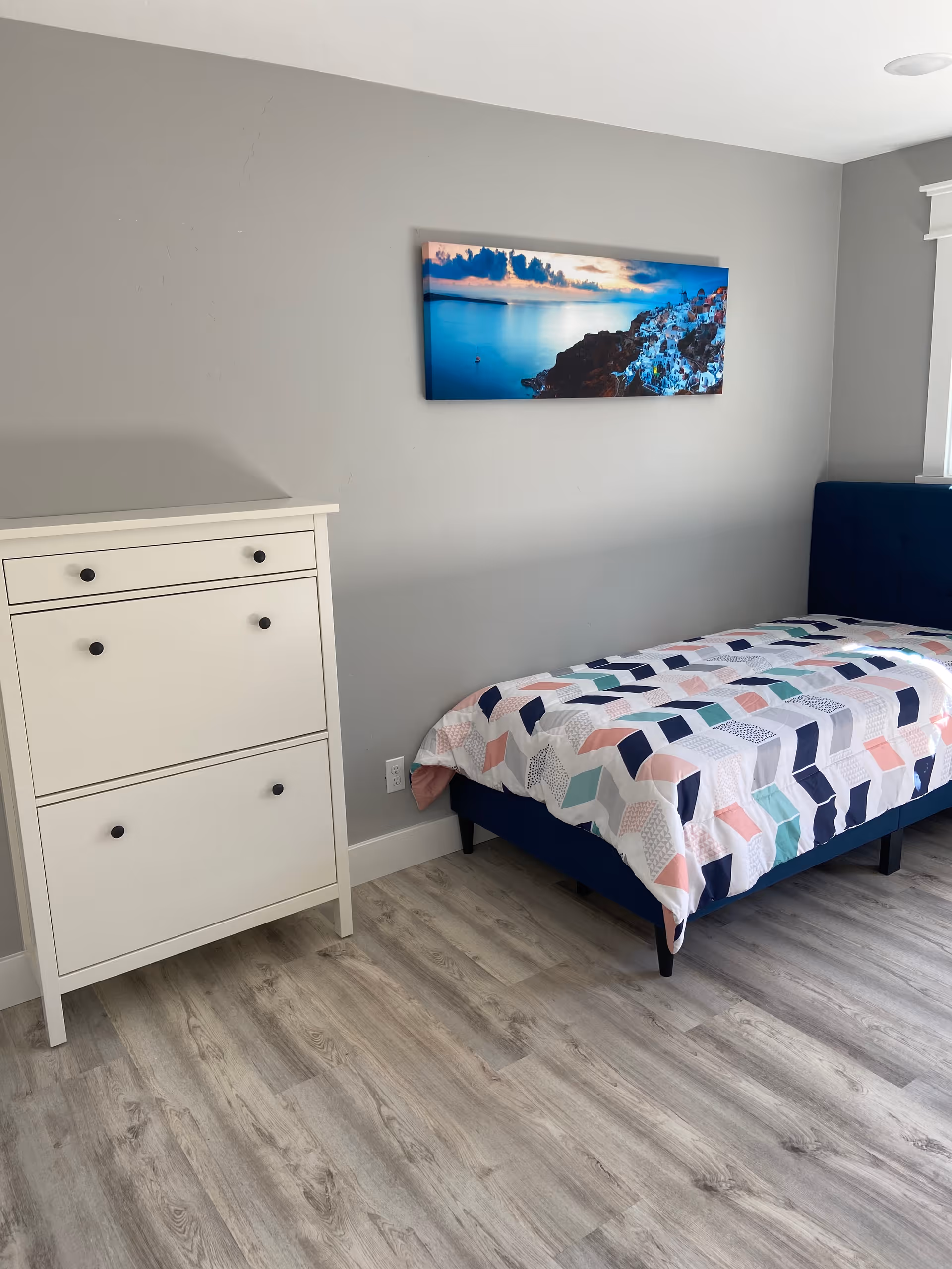 A simple bedroom with a single bed featuring a colorful geometric patterned bedspread. Next to the bed is a white dresser with multiple drawers. The walls are painted light gray, and there is a framed picture of a coastal landscape hanging above the bed. The floor has light wood-style flooring.