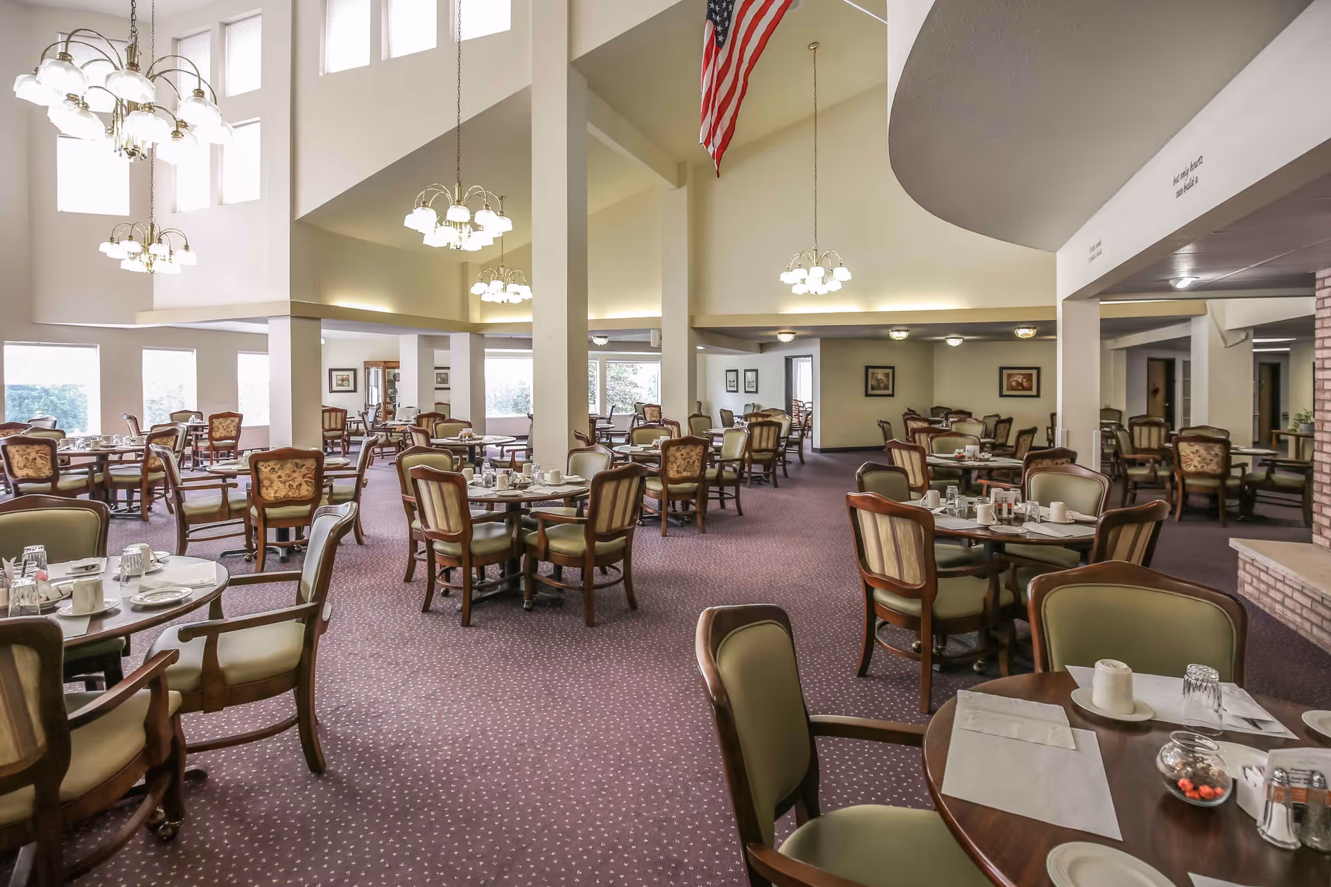 Large dining room with many round tables and upholstered chairs set for service under high ceilings and chandeliers.