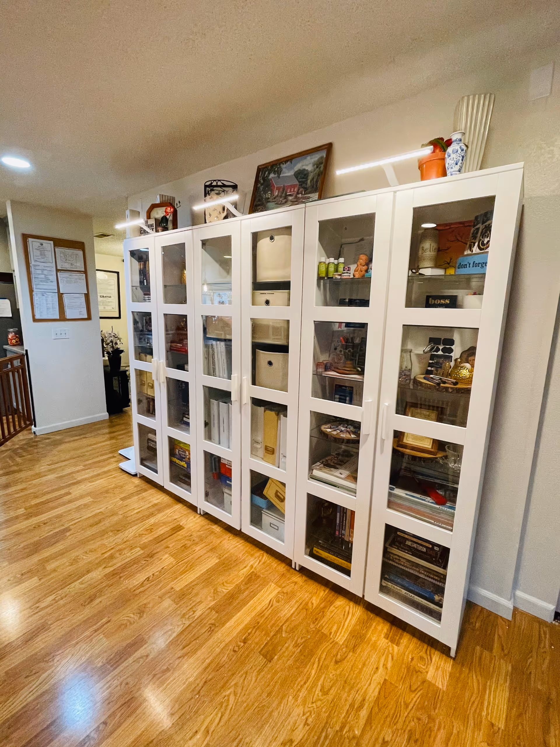 A hallway with wooden flooring and a large white cabinet with glass doors filled with books, boxes, and decorative items. On top of the cabinet are various decorative pieces including a framed painting, a vase, and a small plant. The walls are light-colored and there are framed documents and pictures on the wall to the left.