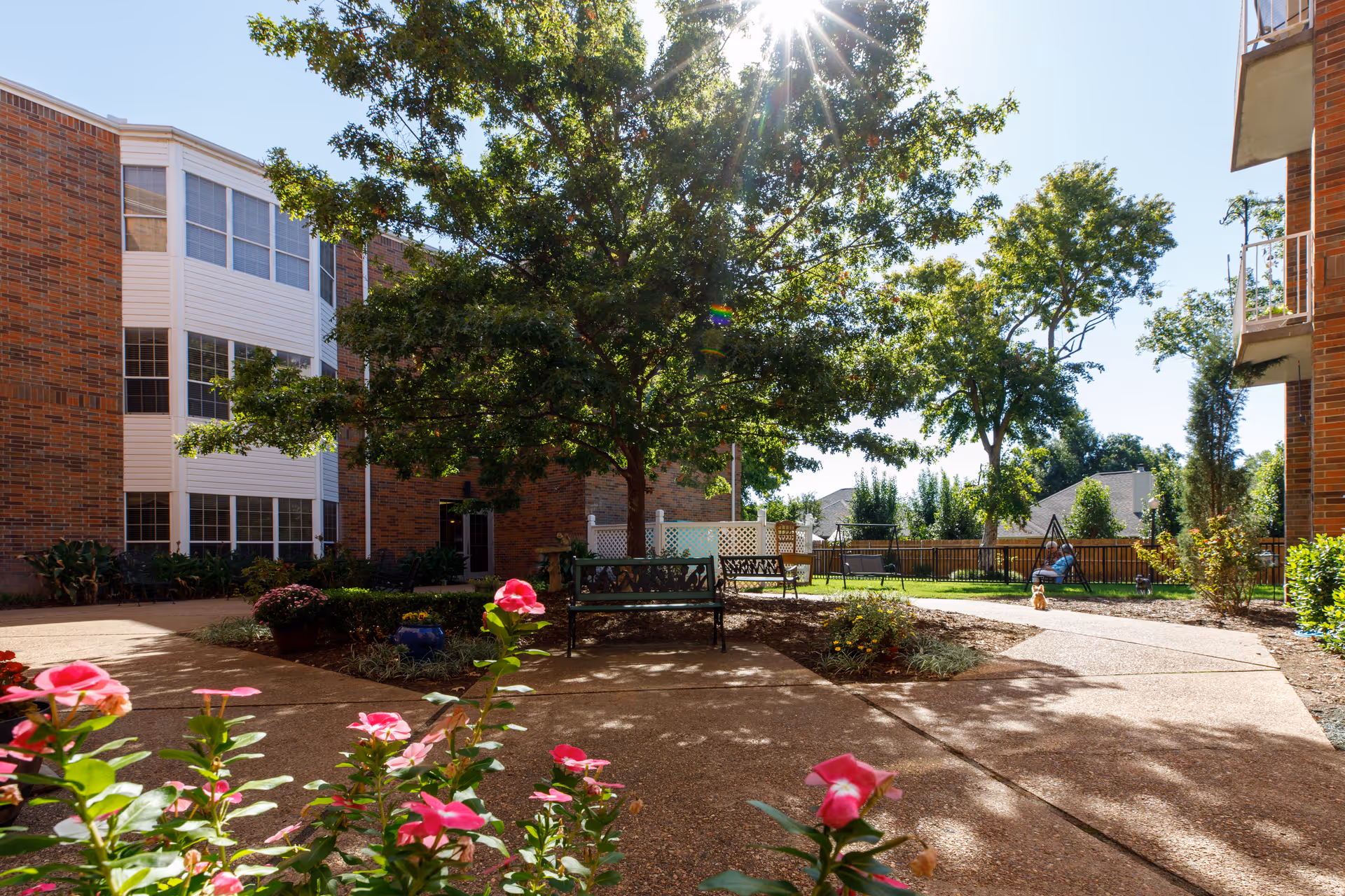 Sunny outdoor courtyard area at Rivermont Independent Living with a large tree providing shade, benches for seating, pink flowers in the foreground, and a swing set in the background. The courtyard is surrounded by brick buildings and has paved walkways.