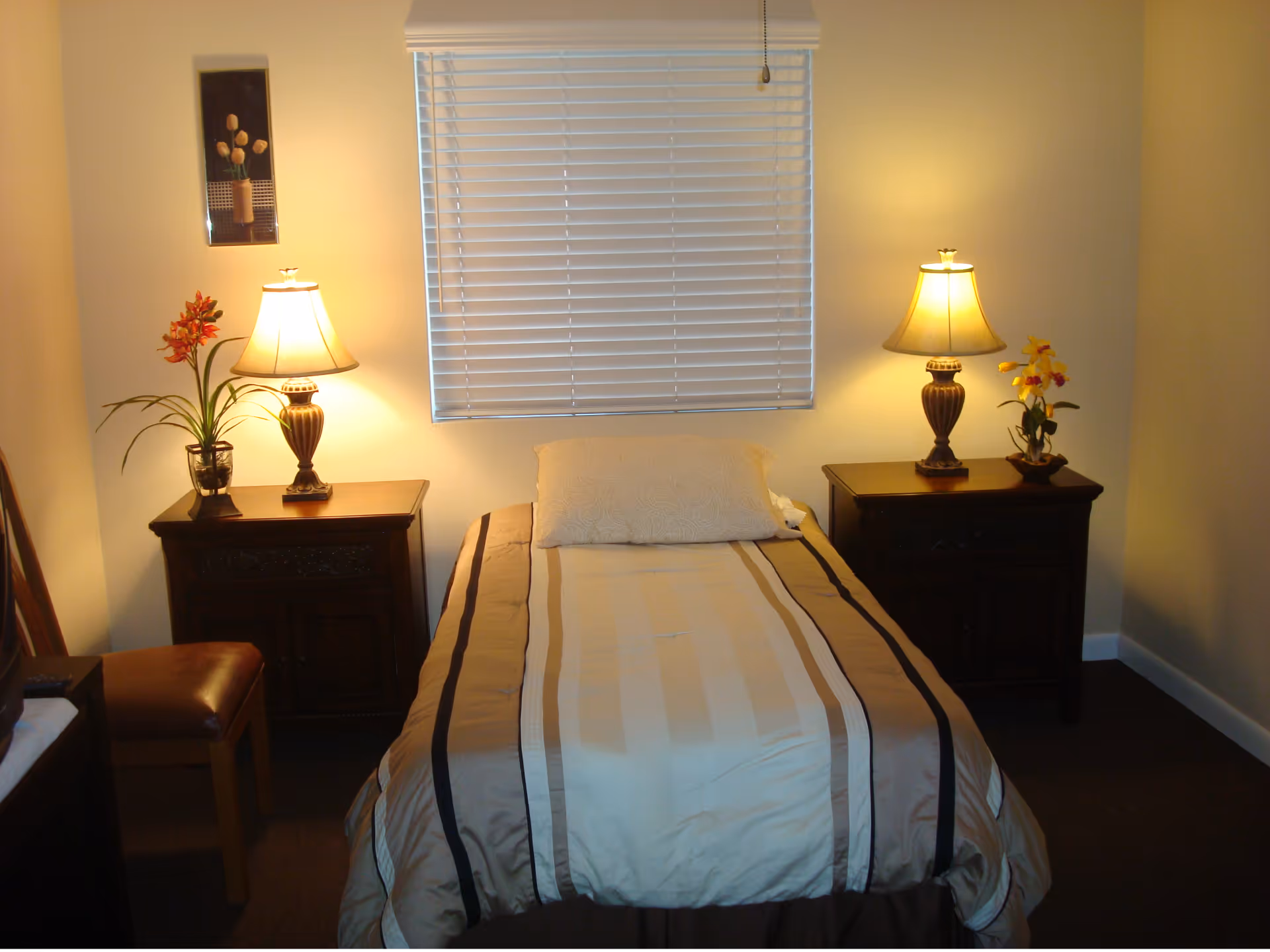 A cozy bedroom with a single bed centered under a window with closed blinds. The bed has a beige and brown striped comforter and a white pillow. On each side of the bed are dark wooden nightstands, each with a table lamp and a potted plant with flowers. A small framed picture hangs on the wall to the left of the window. There is a wooden chair with a brown cushion partially visible on the left side of the room.