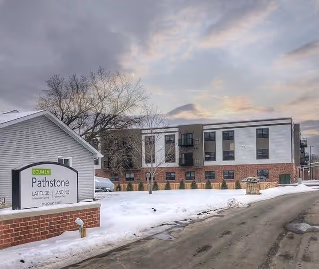 Exterior view of the Ecumen Pathstone senior living facility during winter, showing a three-story building with a mix of brick and siding, a snow-covered lawn, a bare tree, and a sign in front that reads 'Ecumen Pathstone Latitude Landing Memory Care 115 Rogers Street'. The sky is cloudy with some sunlight breaking through.