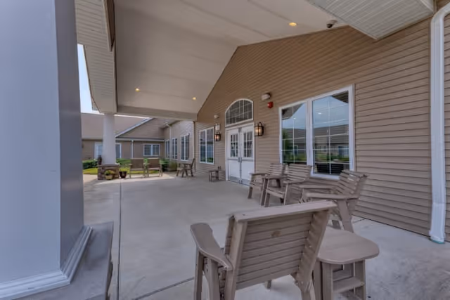 Covered outdoor patio area at Addington Place of Sparta with several wooden chairs and small tables arranged on a concrete floor. The building exterior features beige siding, white trim, and multiple windows. The patio is supported by white columns and has overhead lighting.