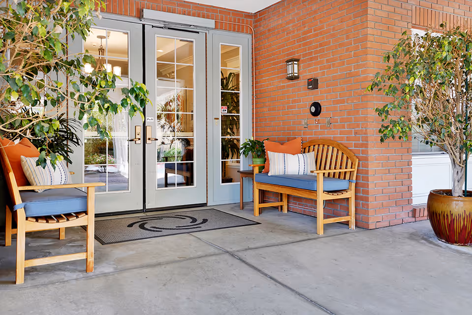 Entrance area of Kirkwood Orange facility with double glass doors, two wooden benches with blue cushions and orange and white pillows, potted plants on either side, and a brick wall background.