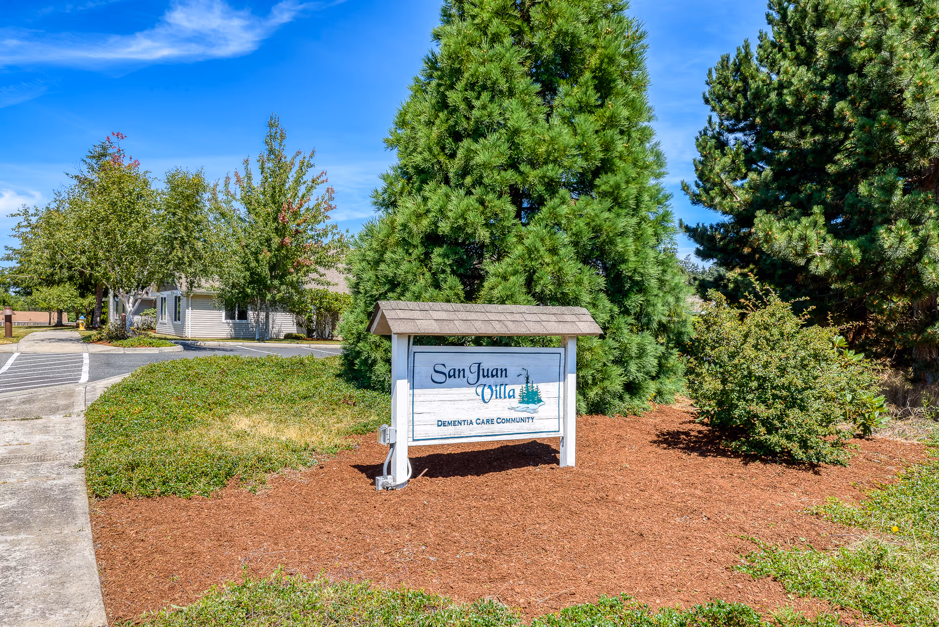 Outdoor view of the entrance area of San Juan Villa Memory Care facility with a white wooden sign displaying the facility name and surrounded by green trees and bushes under a clear blue sky.