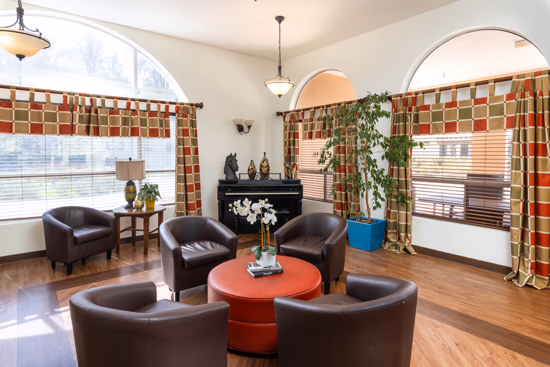 A bright and cozy living room area with four dark brown leather chairs arranged around a round red ottoman. The room features large arched windows with patterned curtains, a small wooden side table with a lamp and plant, a black piano with decorative items on top, and a tall green potted plant in a blue planter. The floor is wooden, and the room is well-lit with natural light.