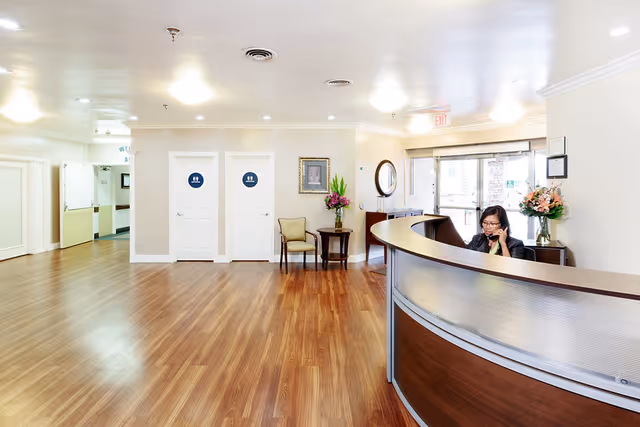 Reception area of Cottonwood Canyon Healthcare Center with a curved wooden reception desk where a woman is seated and talking on the phone. The room has wooden flooring, two white doors with restroom signs, a small table with a chair and a flower vase, and a glass entrance door in the background.