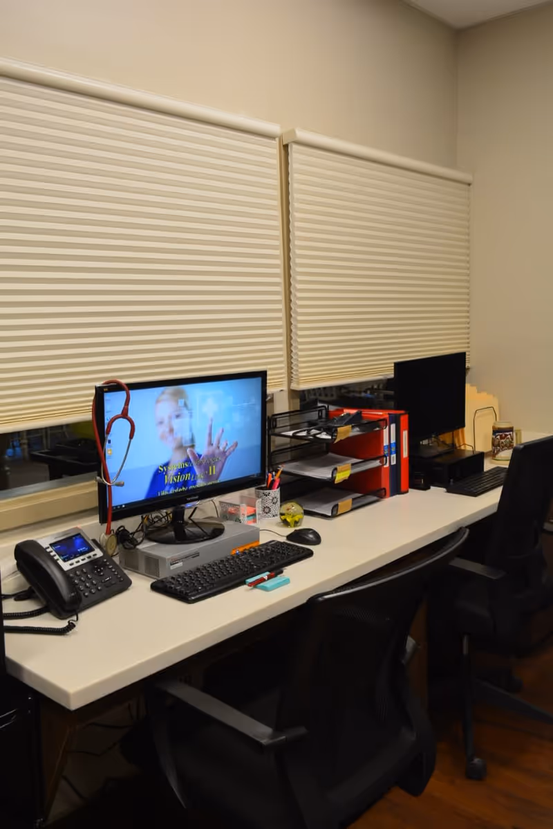 An office workspace with two computer monitors on a long white desk. One monitor displays a medical-themed screen with a stethoscope hanging on its side. The desk also has a telephone, keyboard, mouse, office supplies, and file organizers. Two black office chairs are positioned in front of the desk. The windows behind the desk have closed beige blinds.