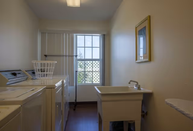 Laundry room with two washing machines, a laundry basket on top, a utility sink with a faucet, an ironing board, a window with vertical blinds, and a framed picture on the wall.