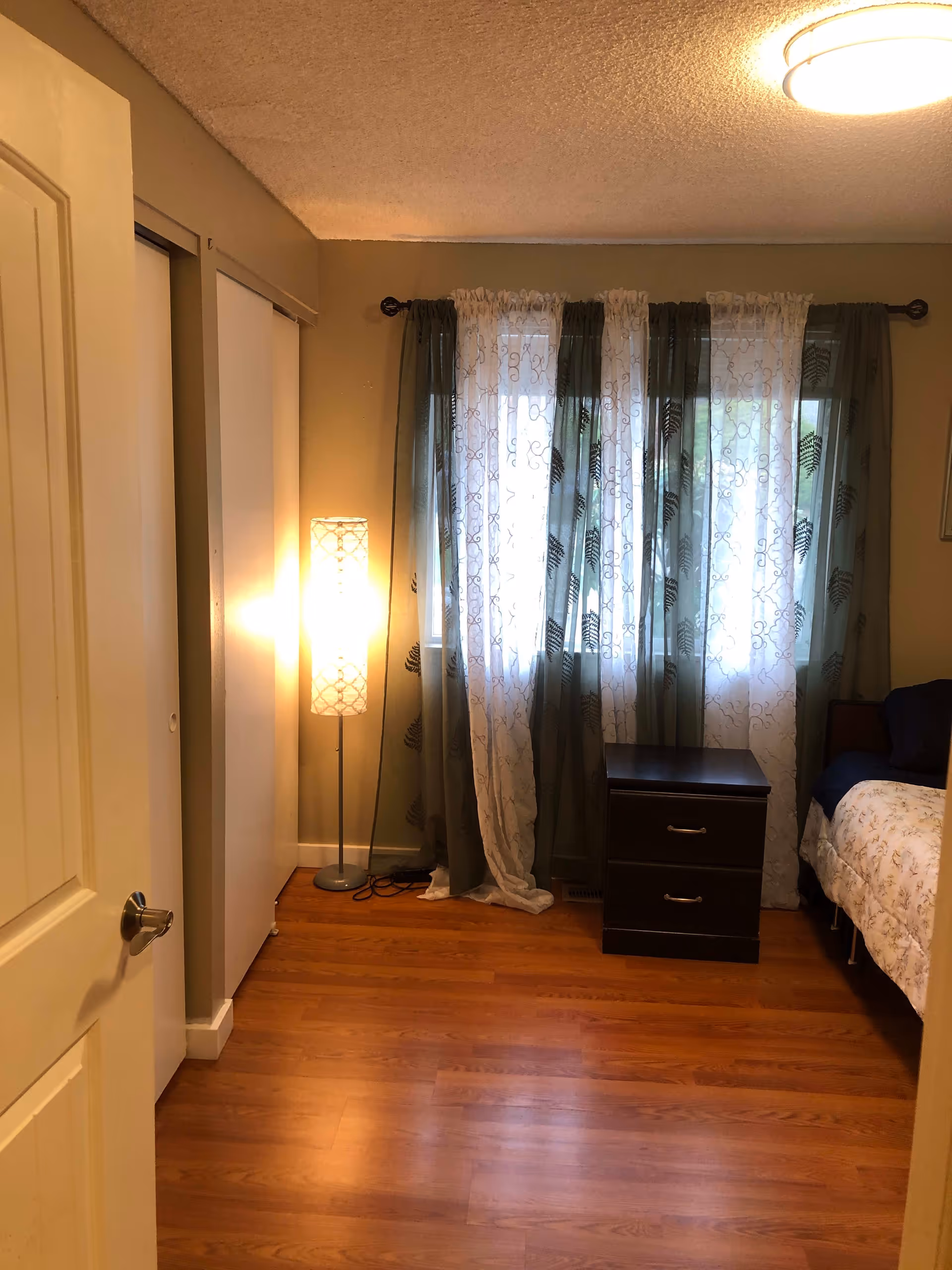 Small bedroom with hardwood floors, a bed and nightstand by a window with patterned curtains and a standing lamp.