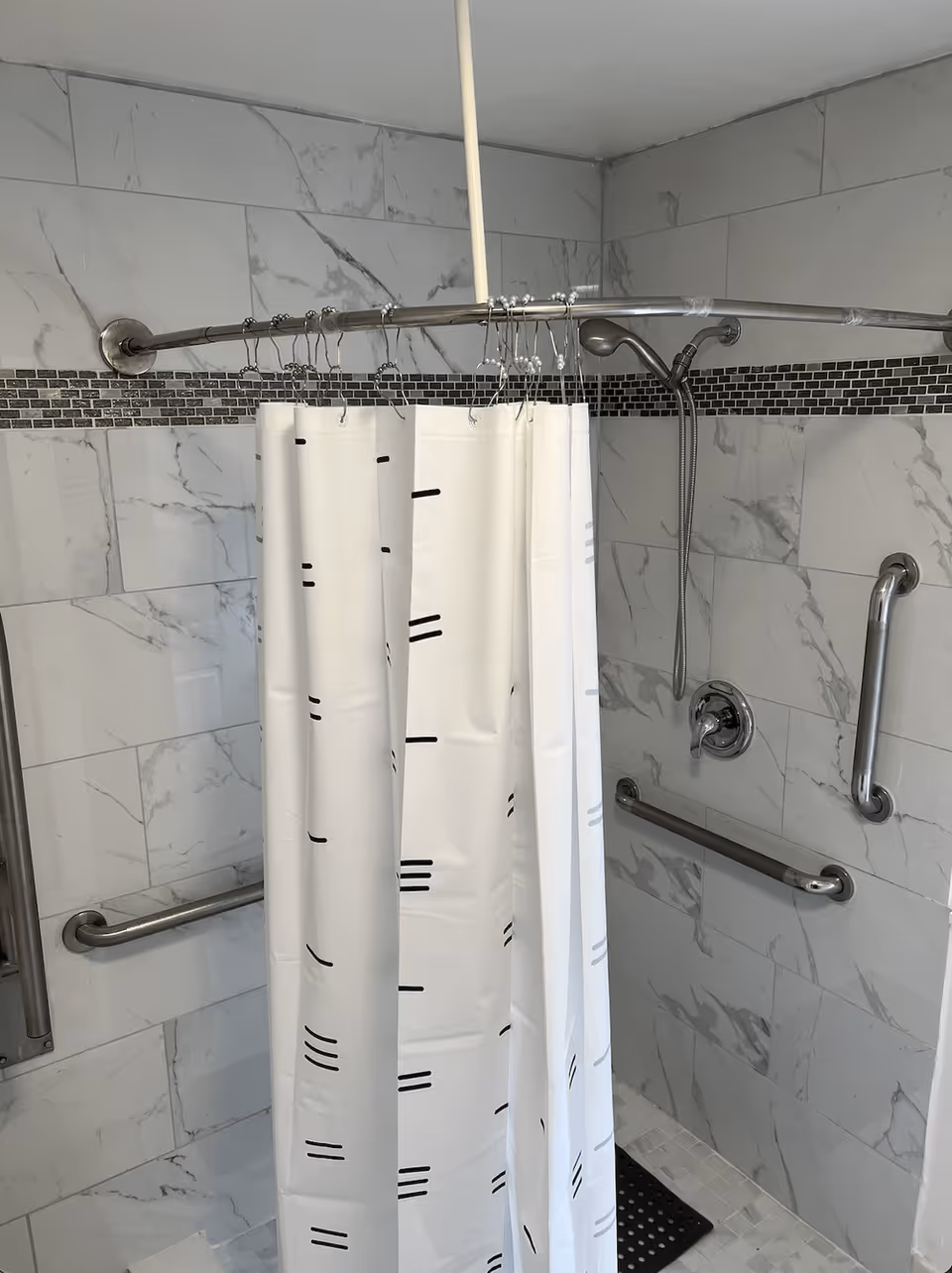 A shower area with white and gray marble-patterned tiles on the walls, a curved metal shower curtain rod with a white shower curtain featuring black lines, a handheld showerhead, and multiple stainless steel grab bars for support.
