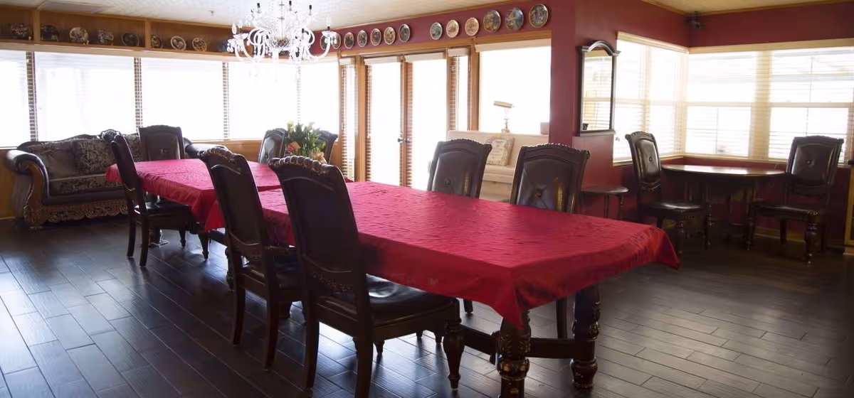 A spacious dining room with a long table covered by a red tablecloth surrounded by dark wooden chairs. The room has large windows with blinds allowing natural light to fill the space. There is a chandelier hanging above the table and decorative plates displayed along the top of the walls. A sofa and additional seating are visible in the background.