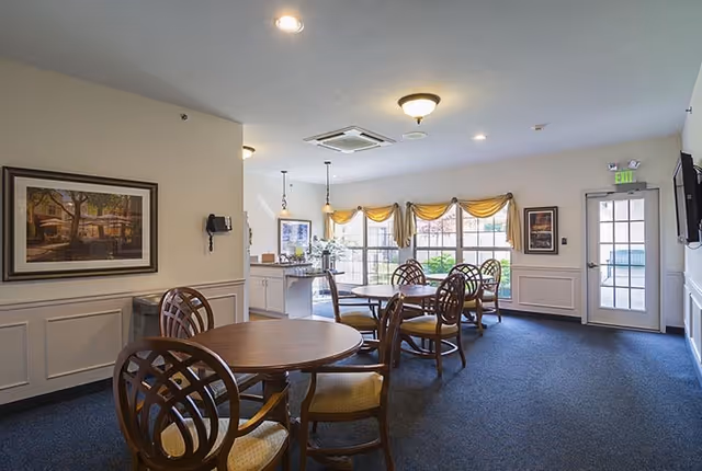 A bright dining area in a senior living facility with round wooden tables and cushioned chairs. Large windows with yellow valances allow natural light to fill the room. There is a door leading outside, framed artwork on the walls, and a small counter area with pendant lights hanging above it.