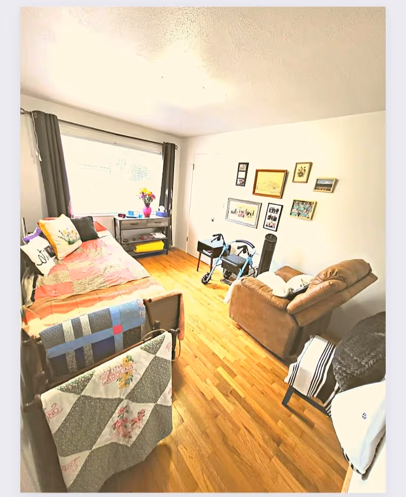 A cozy living room with wooden flooring, a daybed covered with a patchwork quilt and pillows, a small table with a vase of flowers near a large window with dark curtains, a brown recliner chair, two blue walkers, and several framed pictures hanging on a white wall.