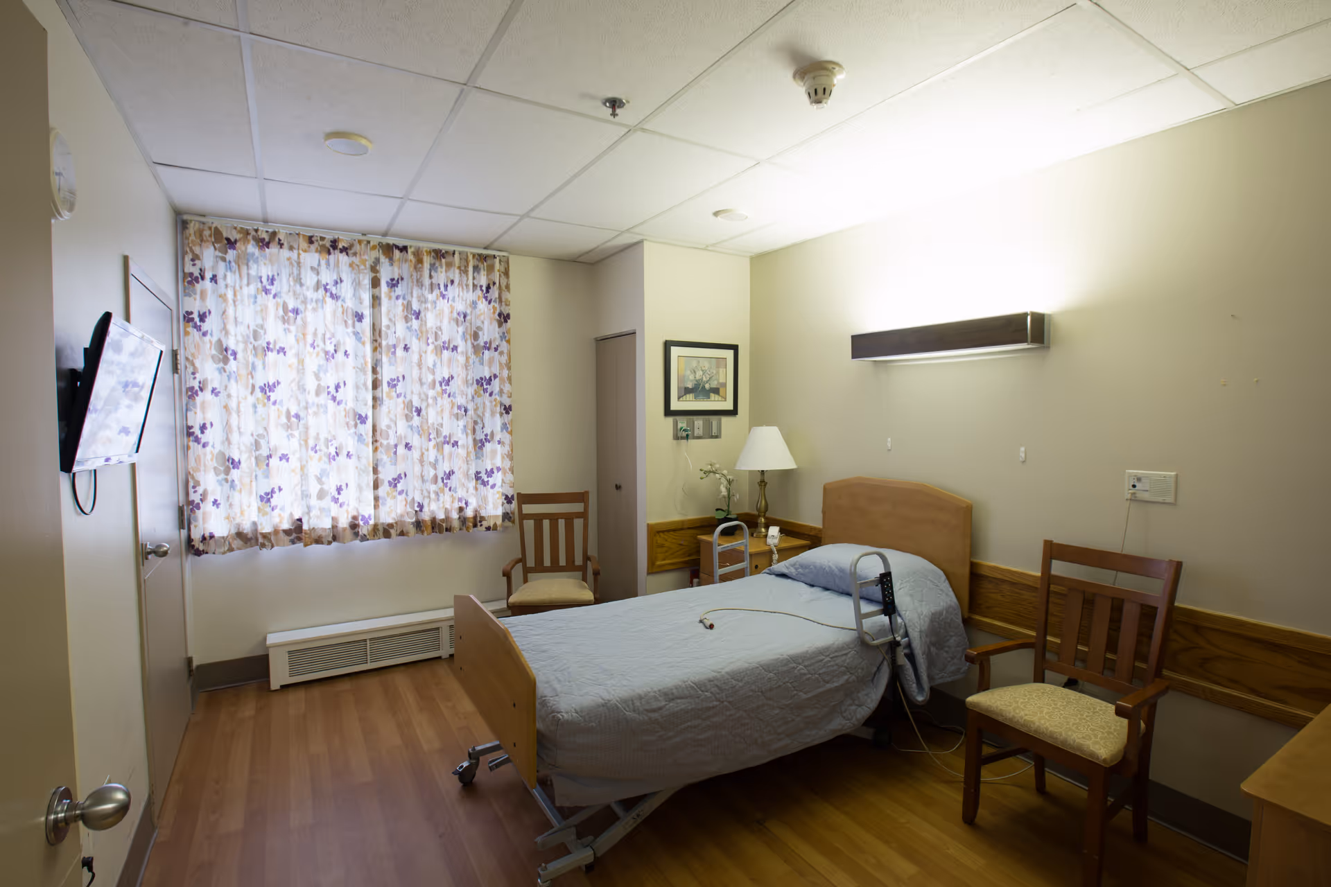 A simple, clean patient room with a single hospital bed covered in a light gray quilt, two wooden chairs with cushions, a small table with a lamp and a framed picture on the wall, floral curtains covering the window, and a wall-mounted TV on the left side.