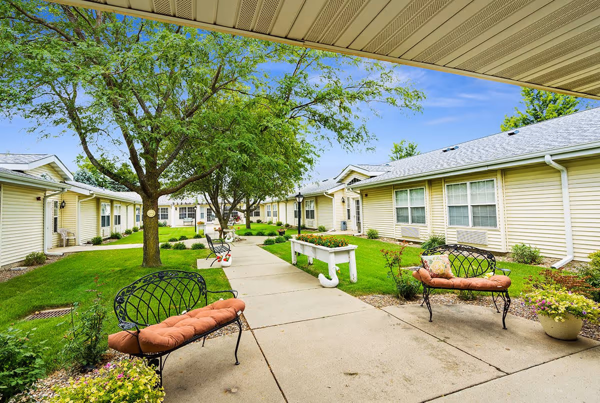 Outdoor courtyard area at Rolling Meadows Senior Living with a concrete walkway, green grass, trees, flower beds, and metal benches with cushions. Single-story beige buildings with white trim surround the courtyard under a blue sky.
