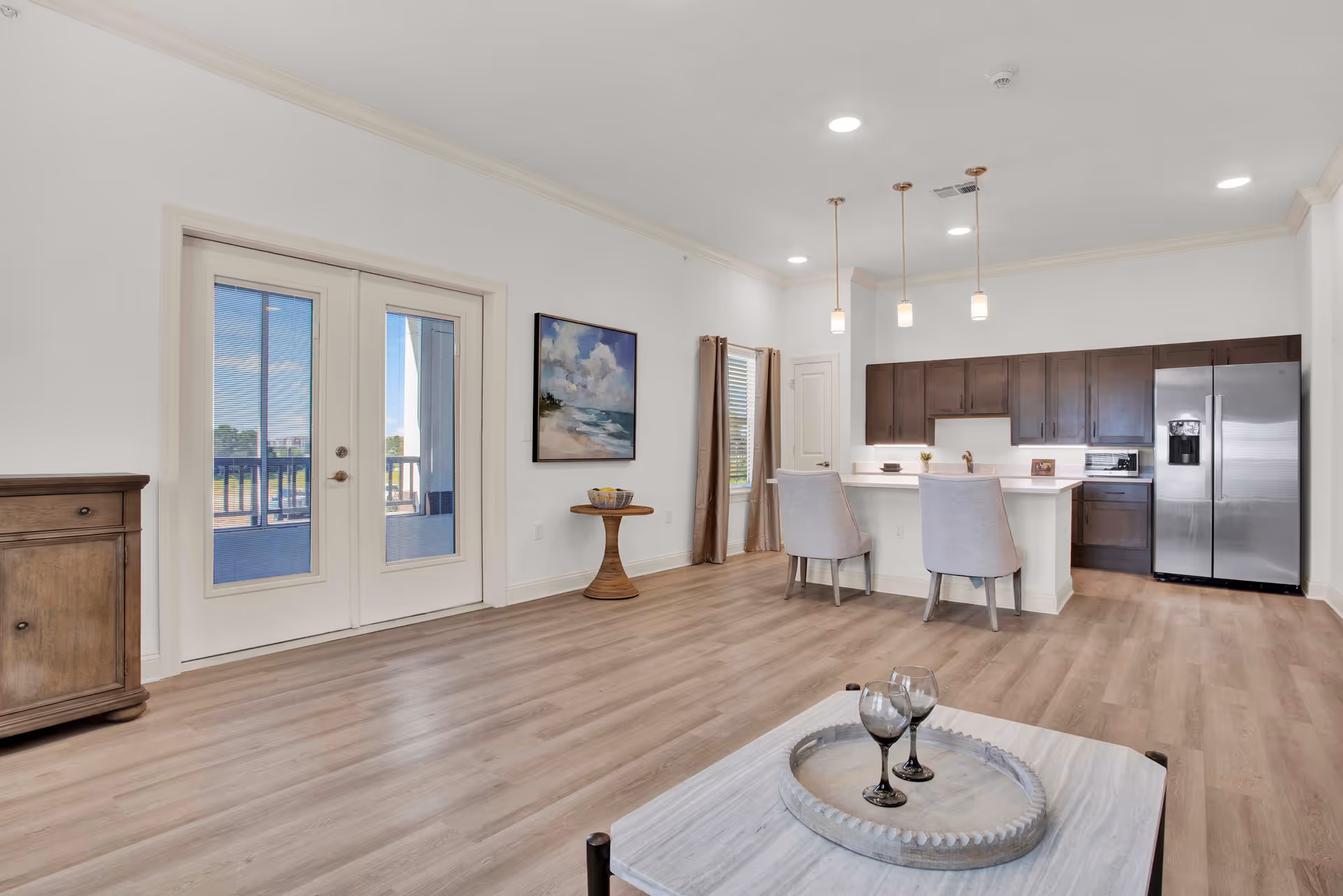 Bright and spacious open-concept living area featuring light wood flooring, a kitchen with dark wood cabinets, stainless steel refrigerator, and a white countertop with two upholstered chairs. There is a small round table with a basket of lemons, a framed beach painting on the wall, and French doors leading to an outdoor balcony.