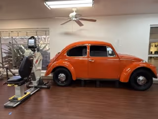 Interior room with wooden flooring featuring an orange vintage Volkswagen Beetle car mounted on a platform. To the left of the car, there is exercise equipment including a recumbent bike. The room has large windows with blinds, a ceiling fan, and fluorescent lighting.