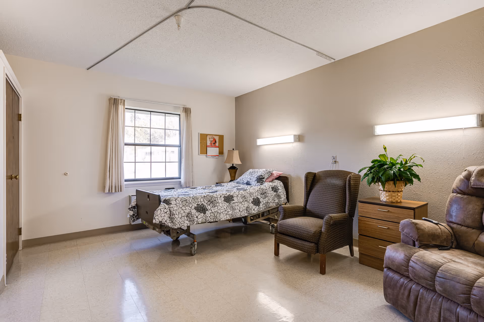 A senior living facility bedroom with a hospital-style bed covered in a floral bedspread, a window with beige curtains, a wooden nightstand with a lamp and a potted plant, a cushioned armchair, and a recliner chair. The room has beige walls and a tiled floor.