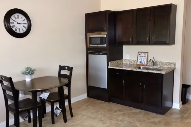 Small kitchenette with dark wood cabinets, a granite countertop, mini fridge and microwave, and a round dining table with two chairs beneath a wall clock.