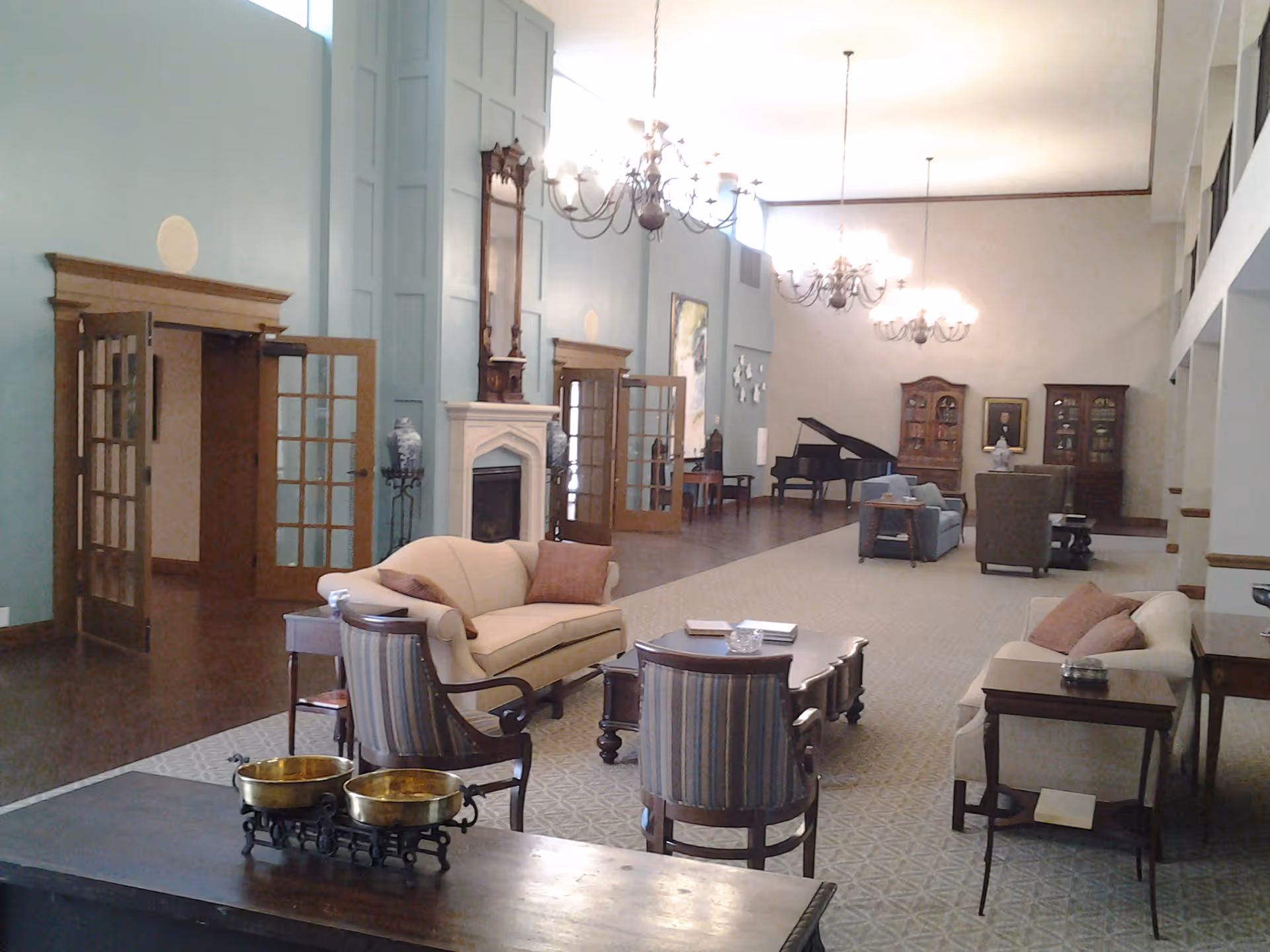 A spacious and elegant living room area with high ceilings, chandeliers, and large windows. The room features multiple seating arrangements including sofas and armchairs, a fireplace with a tall clock above it, a grand piano in the background, and wooden cabinets along the far wall. The decor is classic and inviting, with soft lighting and a patterned carpet.