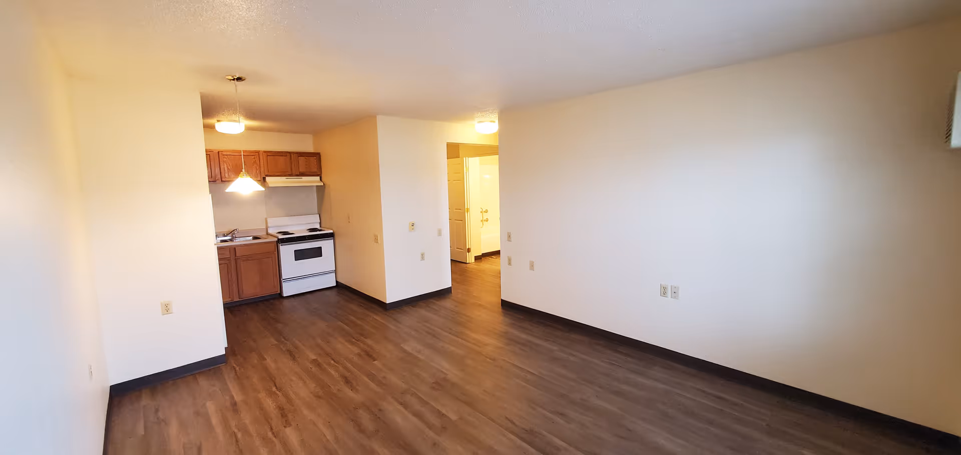 Empty apartment interior with wood flooring, white walls, and a small kitchen area featuring wooden cabinets, a white stove, and a hanging light fixture. There is an open doorway leading to another room or hallway.