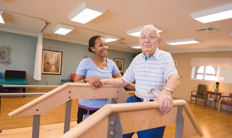 An elderly man using parallel bars for physical therapy in a rehabilitation center, assisted by a smiling female caregiver. The room has wooden flooring, framed artwork on the walls, and chairs near a window with blinds.