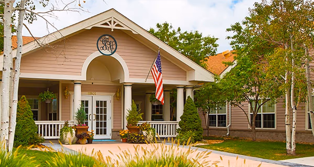 Front exterior view of a senior living facility building with a covered entrance supported by white columns, an American flag hanging near the entrance, potted plants, trees, and a well-maintained lawn.