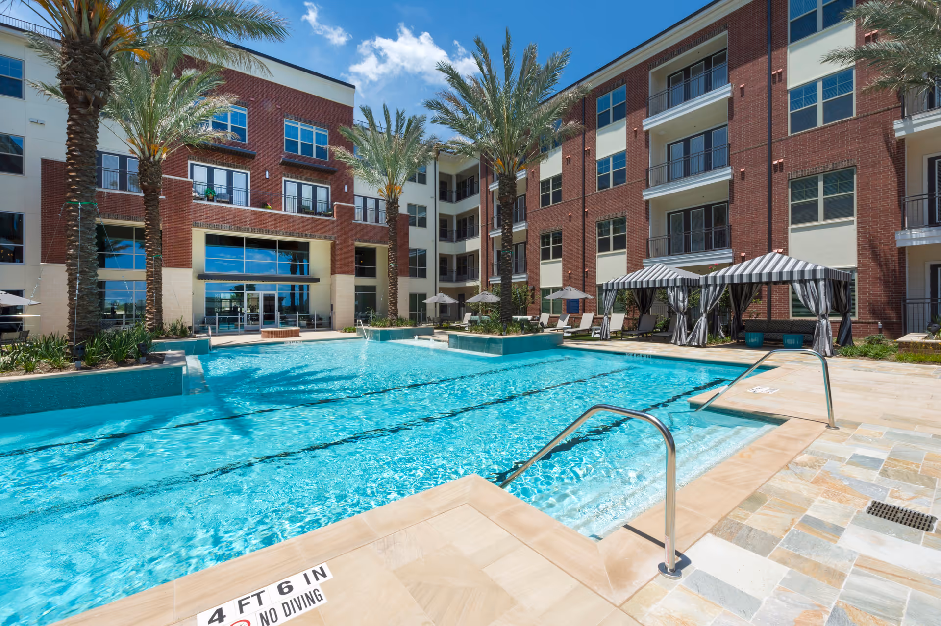 Outdoor swimming pool area at Overture Sugar Land with clear blue water, surrounded by palm trees, lounge chairs, umbrellas, and striped cabanas. The pool is adjacent to a multi-story brick and cream-colored building under a bright blue sky with some clouds.