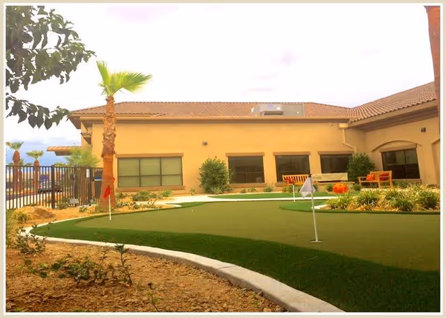 Outdoor putting green area with small flags, surrounded by landscaping with bushes and palm trees, adjacent to a beige building with multiple windows and benches.