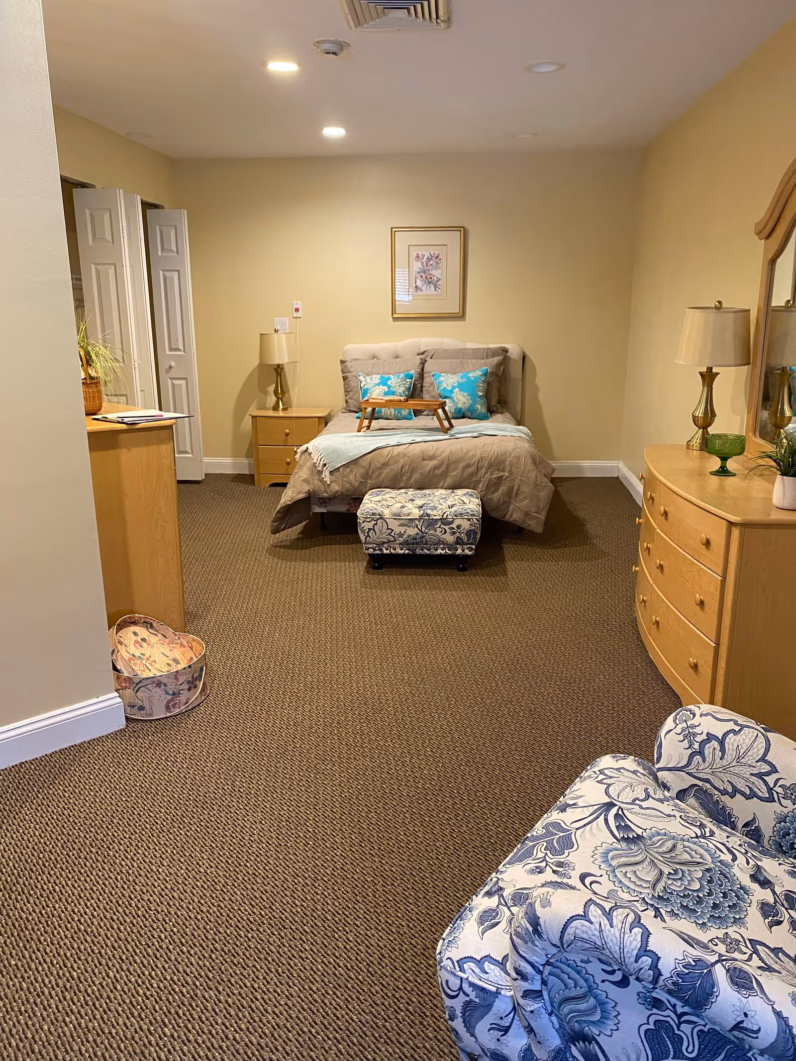 A cozy bedroom with a bed centered against a beige wall, decorated with two blue floral pillows and a light blue throw blanket. There is a small wooden tray on the bed. To the right, there is a wooden dresser with a mirror, a lamp, and some decorative items. A blue and white floral armchair is partially visible in the foreground. The room has brown carpet flooring and soft overhead lighting.