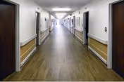 A long, clean hallway in a healthcare or senior living facility with wooden doors on both sides, handrails along the walls, and bright overhead lighting.
