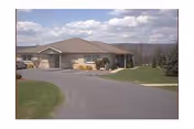 Single-story senior living facility building with a curved driveway, lawn and shrubs under a partly cloudy sky.