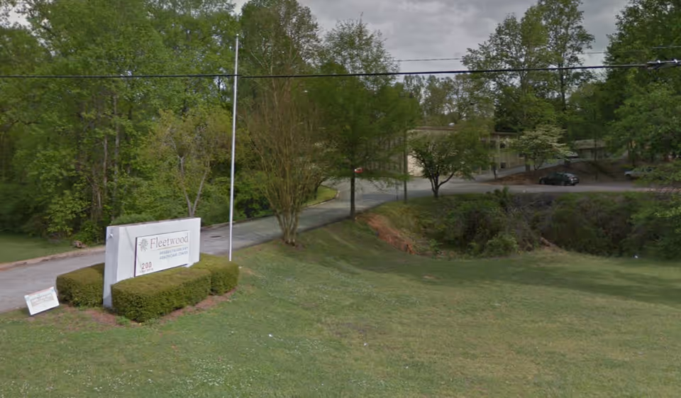 Outdoor view of Fleetwood Post-Acute facility entrance with a sign displaying the facility name and address, surrounded by green grass, bushes, and trees under a cloudy sky.