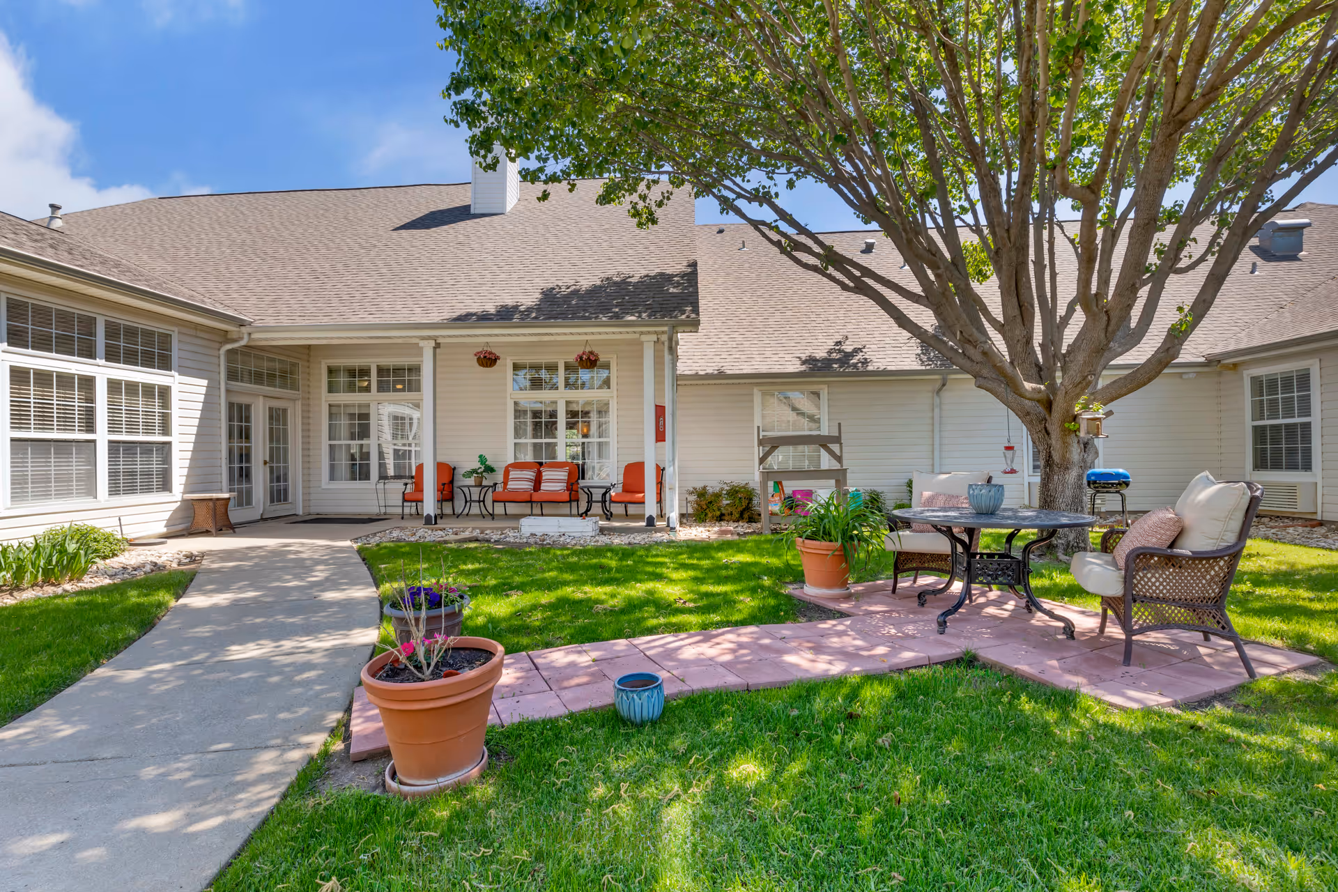 Outdoor patio area at Brookdale Denton North featuring a large tree, green grass, potted plants, and a seating area with cushioned chairs and a round table. The building exterior is light-colored with multiple windows and a covered porch with additional seating.