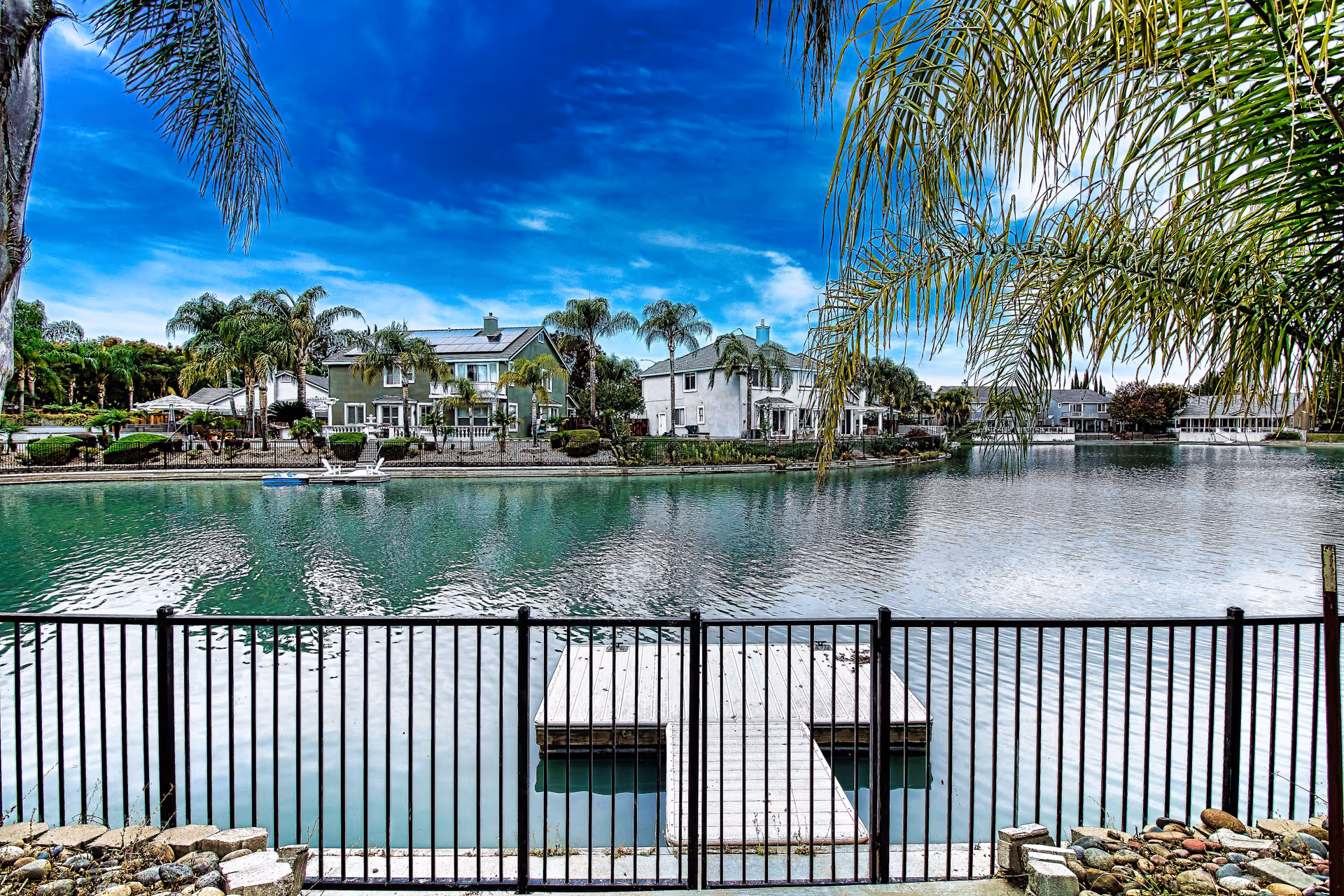 View of a serene lake with a small dock behind a black metal fence, surrounded by palm trees and residential houses under a bright blue sky with some clouds.
